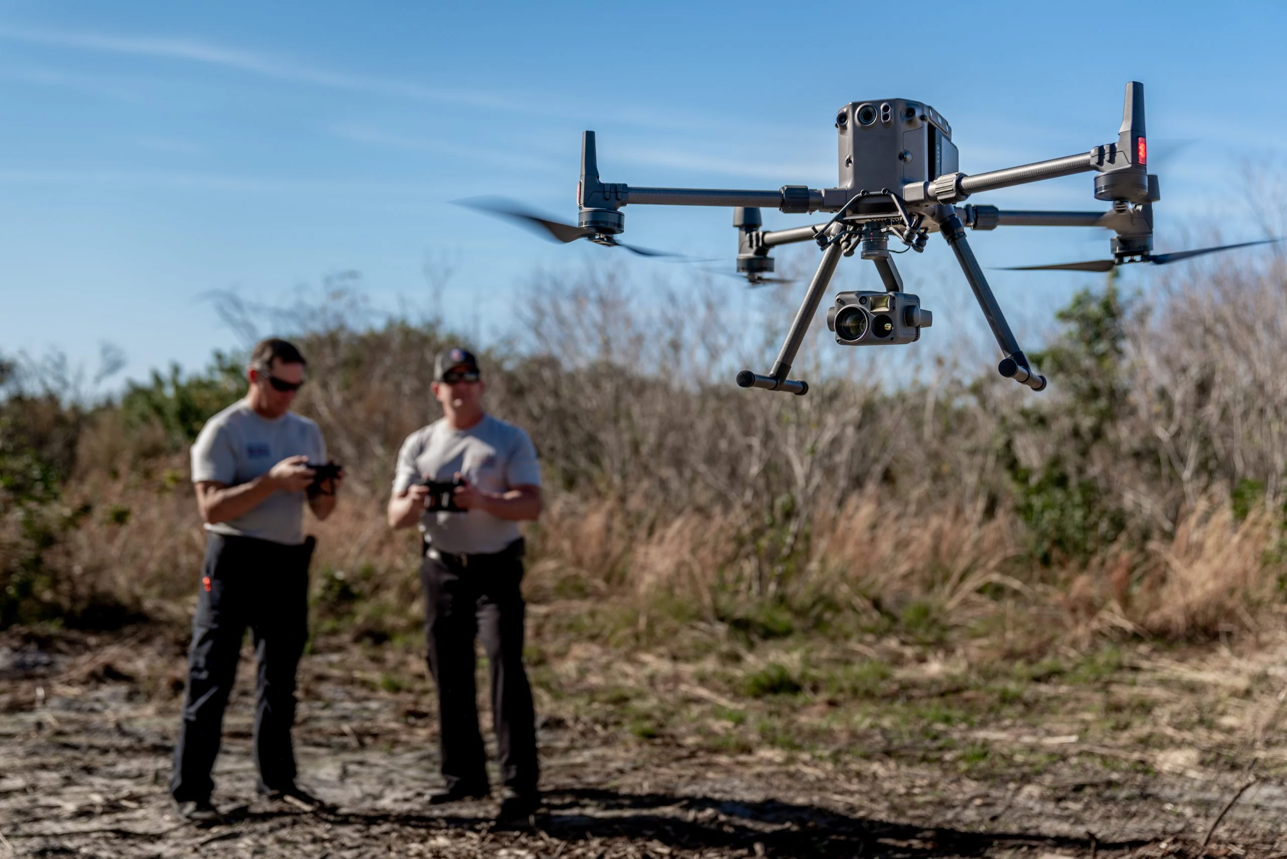 due uomini in un campo, con i controller di un drone, e in primo piano un drone con telecamera che si libra in aria